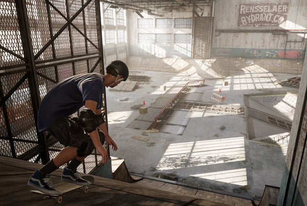 Skateboarder wearing a helmet crouches at the edge of a ramp inside an indoor skatepark, preparing to drop in, with rails, ramps, and sunlight streaming through large windows.