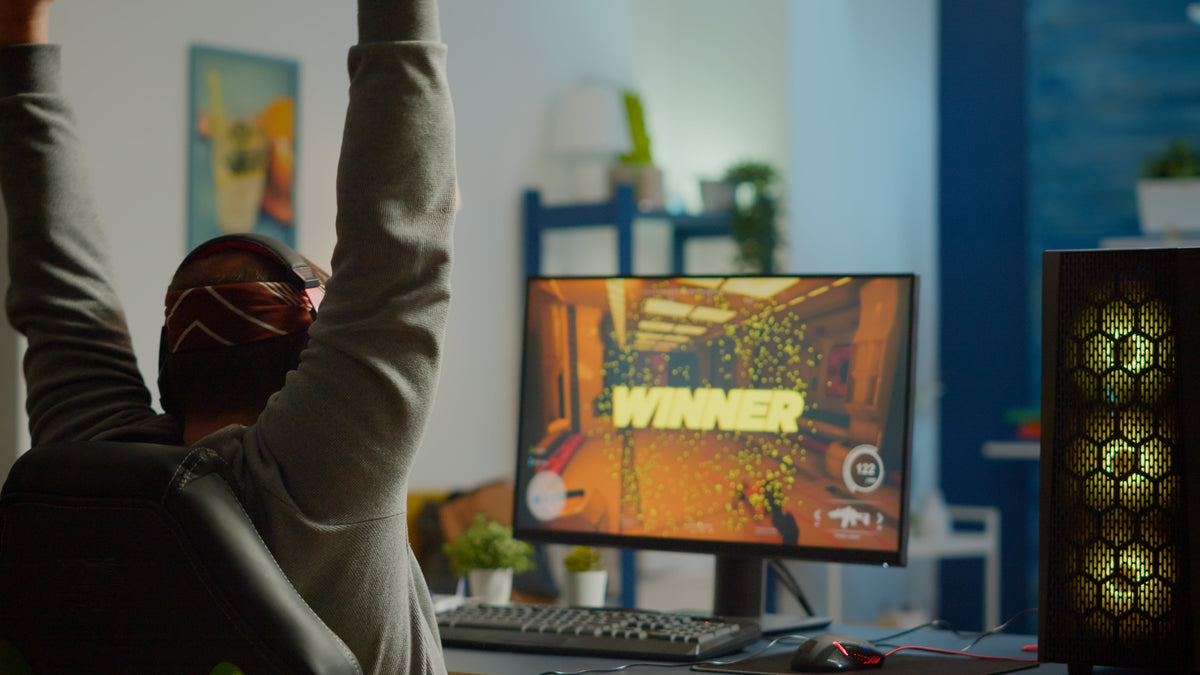 Gamer raising their arms in victory while sitting at a desk, with a PC monitor displaying the word Winner in a brightly lit game scene.