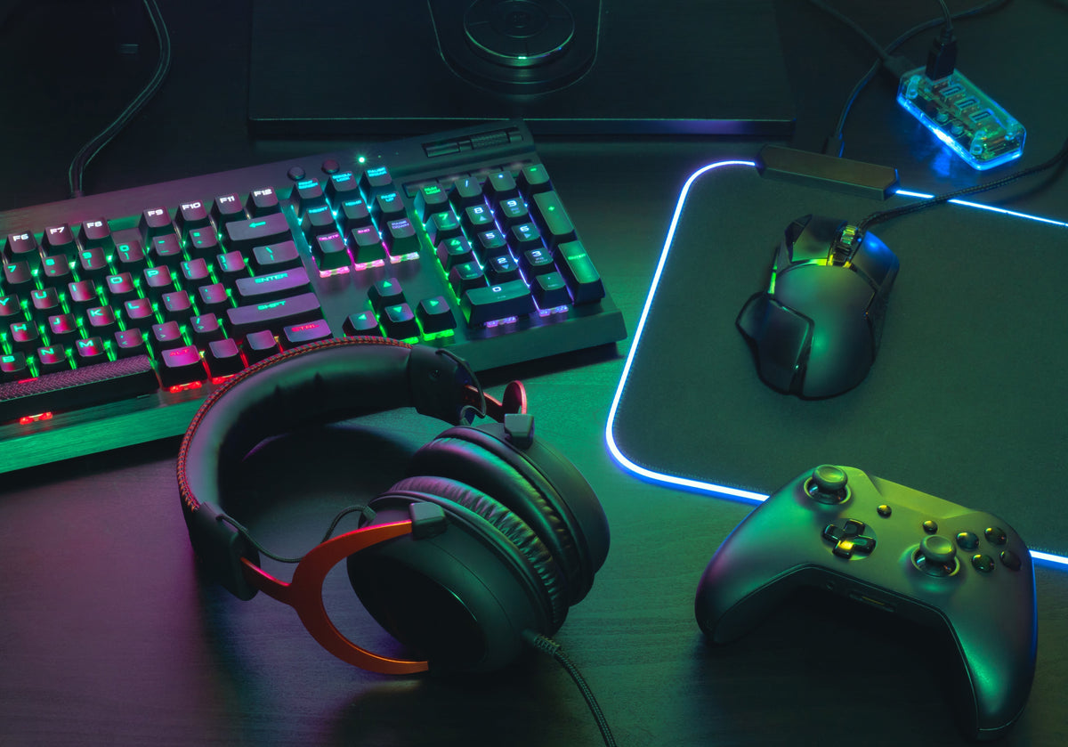 A gaming setup featuring a mechanical RGB keyboard, an illuminated gaming mouse on an LED mousepad, a wireless controller, and over-ear headphones with red accents, all arranged on a dark desk under colorful ambient lighting.