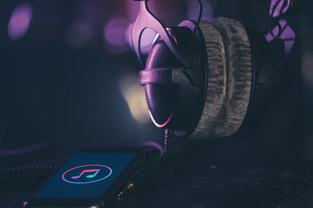 A pair of over ear headphones rests next to a smartphone displaying a music note icon on its screen. The scene is dimly lit with soft purple and blue lighting, creating a moody, atmospheric focus on music and audio.