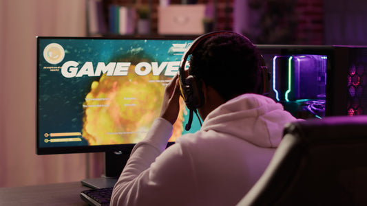 A gamer wearing a headset sits at a desk facing a monitor that displays a “Game Over” screen, resting their hand on their head in frustration while a colorful PC setup glows beside the screen.
