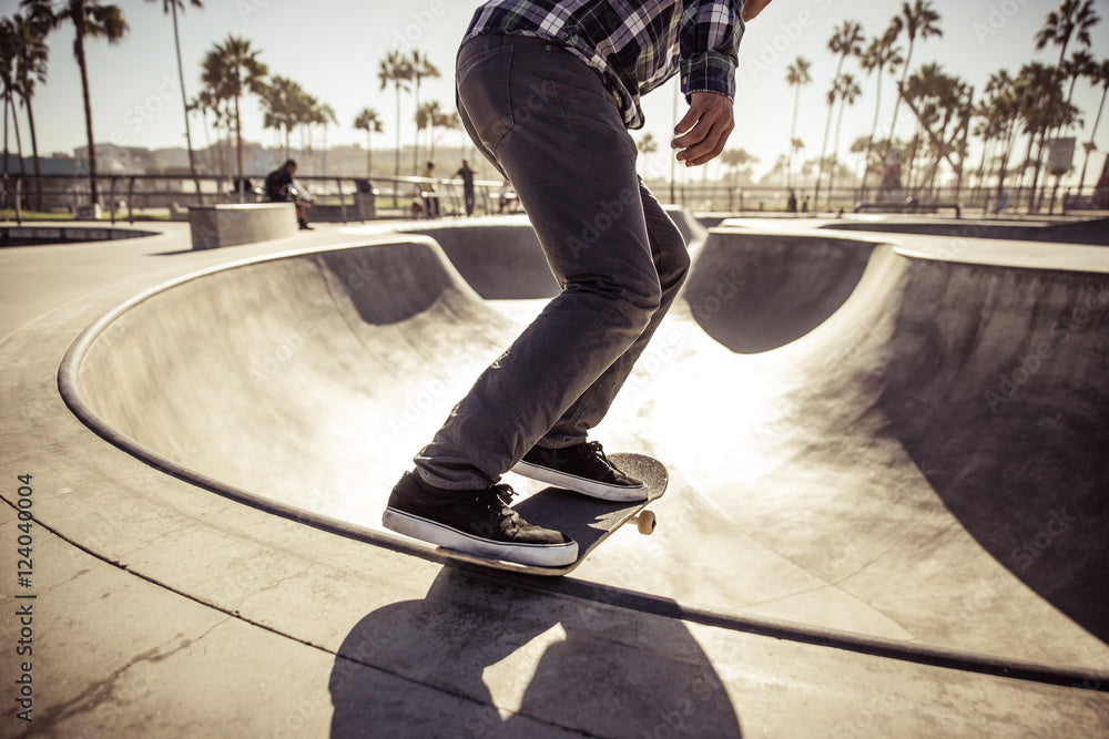 Skateboarder riding along the edge of a concrete skate bowl at an outdoor skate park with palm trees in the background during a sunny day.