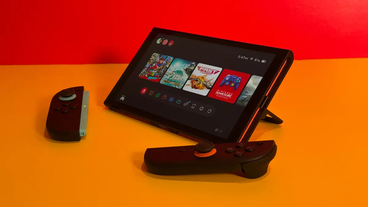 A Nintendo Switch 2 in tabletop mode with detached controllers on an orange surface, displaying the home screen with several game titles.